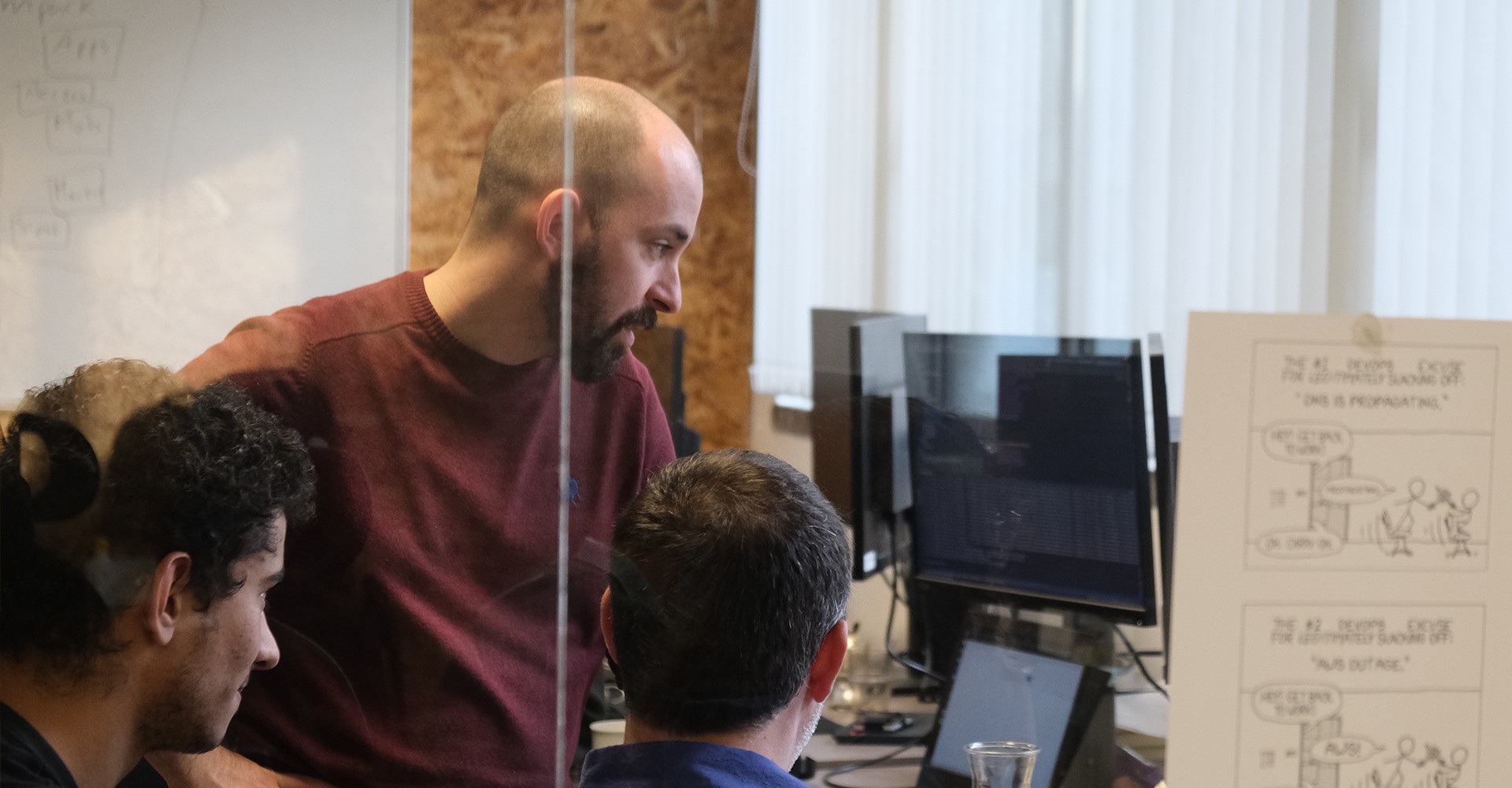 Three young men look focused at a computer screen while solving a programming problem in an office full of computers.