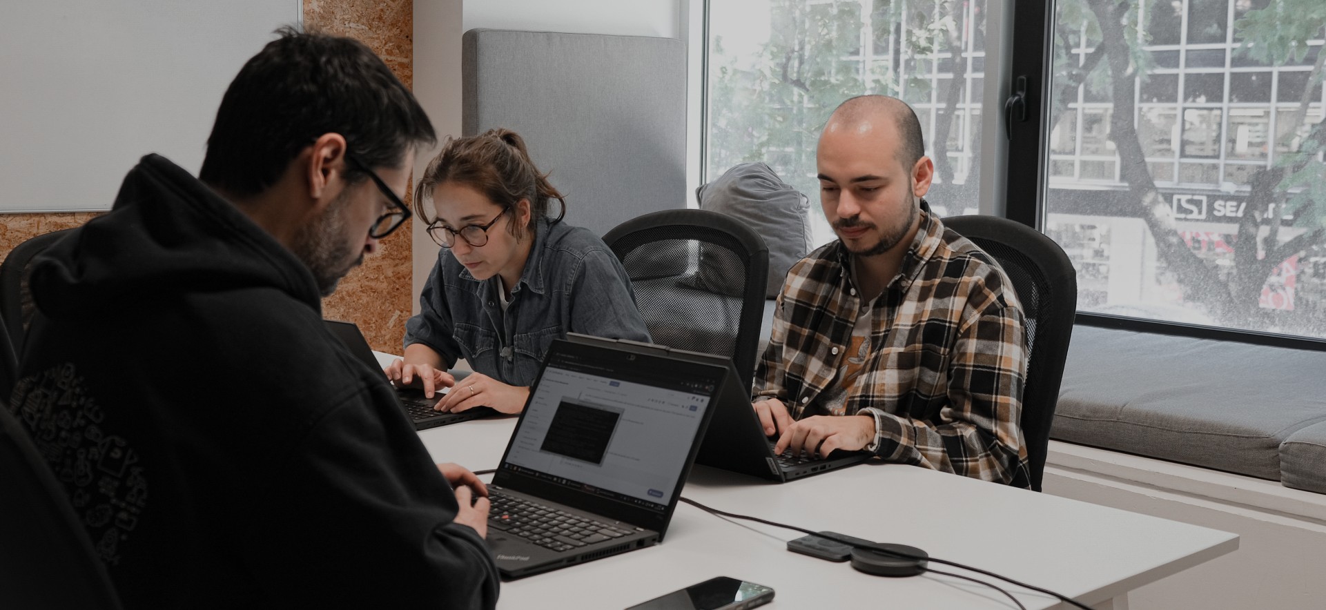 Three young people look focused at a computer screen while solving a programming problem in an office full of computers.